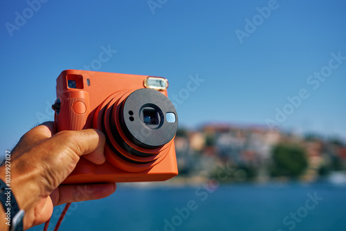 Close-up of tourist hand holding an orange instant camera in retro style with coastal town and blue sea in background. Summer travel concept. Equipment for capturing moments photo