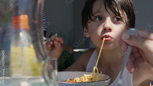 Young boy slurping a long noodle while eating spaghetti, using a fork to twirl the pasta and enjoying his meal outdoors at a table during the day photo