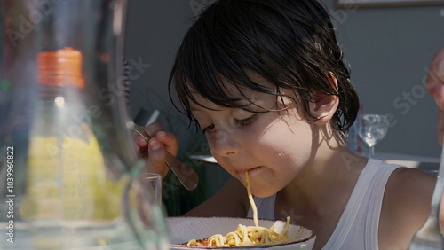 Young boy slurping a long noodle while eating spaghetti, using a fork to twirl the pasta and enjoying his meal outdoors at a table during the day photo