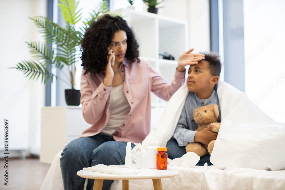 African descent mother checks temperature of sick young boy at home. Boy lies on bed holding teddy bear, while mother consults doctor by phone. Medical supplies visible nearby. African descent mother checks temperature of sick young boy at home. Boy lies on bed holding teddy bear, while mother consults doctor by phone. Medical supplies visible nearby.