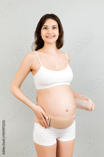 Portrait of pregnant woman in underwear dressing elastic maternity band at gray background with copy space. Orthopedic abdominal support belt concept