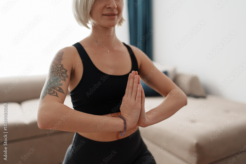A woman in a black tank top meditates with hands in prayer position, embodying calm and mindfulness. She is practicing yoga at home in a serene setting, promoting wellness and tranquility. A woman in a black tank top meditates with hands in prayer position, embodying calm and mindfulness. She is practicing yoga at home in a serene setting, promoting wellness and tranquility.