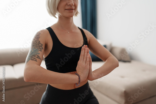 A woman in a black tank top meditates with hands in prayer position, embodying calm and mindfulness. She is practicing yoga at home in a serene setting, promoting wellness and tranquility.