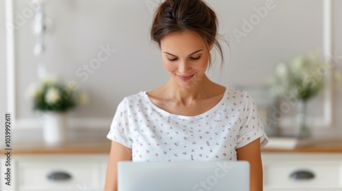 A young woman smiles while working on a laptop in a bright, modern kitchen, emphasizing a cozy and productive home environment.