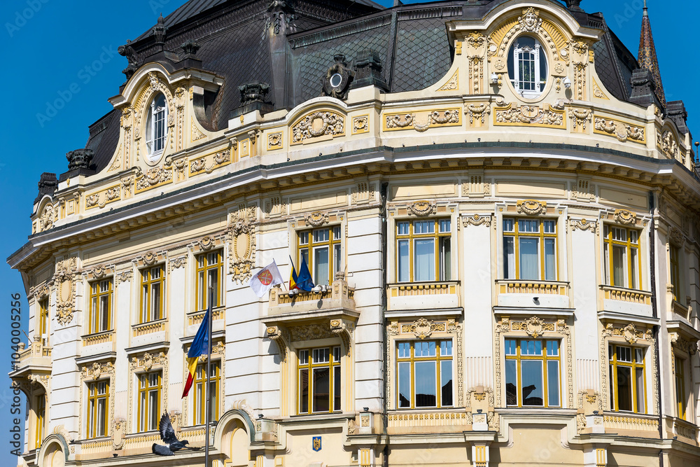 A close-up of a historic building facade in Sibiu, Romnania, featuring a curved window surrounded by intricate decorative elements. The building has a dark roof and a light-colored exterior. A close-up of a historic building facade in Sibiu, Romnania, featuring a curved window surrounded by intricate decorative elements. The building has a dark roof and a light-colored exterior.