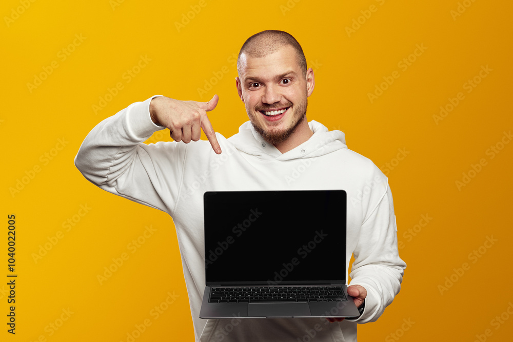 Trendy hipster guy with beard wearing white hoodie smiling and pointing at new laptop in hands, isolated over yellow background. Trendy hipster guy with beard wearing white hoodie smiling and pointing at new laptop in hands, isolated over yellow background.