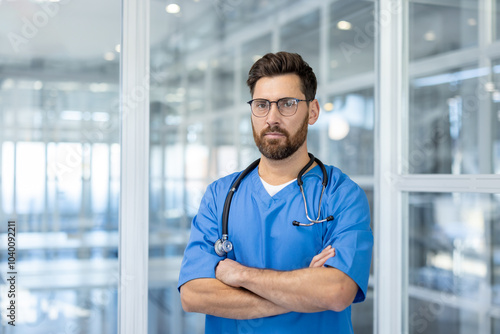 Confident male doctor in blue scrubs with stethoscope, standing in modern medical facility. Medical professional with glasses portrays expertise and trust in healthcare setting.