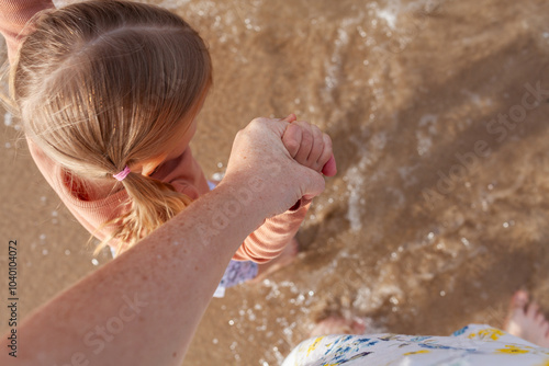 Parents pov looking down on mums hand holding child's as they wade into sandy ocean water