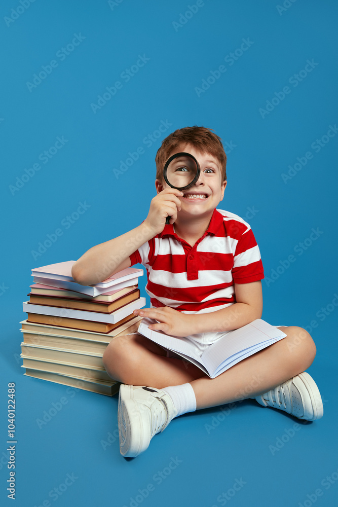 Vertical photo of excited smart boy looking at camera through magnifying glass and holding open book while sitting crossed legged on floor near bunch of books, isolated over blue background Vertical photo of excited smart boy looking at camera through magnifying glass and holding open book while sitting crossed legged on floor near bunch of books, isolated over blue background
