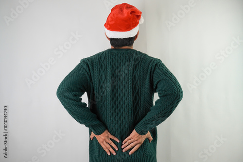 Young asian man wearing santa clause hat and green long sleeved sweater is suffering lower back muscle pain with grimacing face, isolated white background. Concept for Christmas and New Year Party