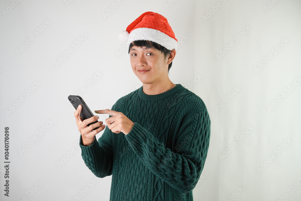 Excited young asian man wearing santa clause hat and green long sleeved sweater is using smartphone, isolated over white background. Concept for Christmas Holiday and New Year Party Excited young asian man wearing santa clause hat and green long sleeved sweater is using smartphone, isolated over white background. Concept for Christmas Holiday and New Year Party