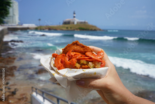 Obraz na płótnie Acaraje traditional dish and street food from Northeast Brazil with Farol da Barra Lighthouse and Bay of All Saints on the background, Salvador de Bahia, Brazil w salonie