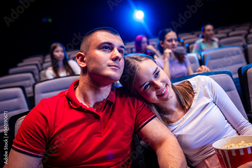 A young couple is watching a movie in the cinema. She lays her head on his shoulder and they eat popcorn.