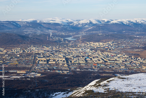 View from the mountain to the city of Magadan. Top view of city blocks, streets and buildings. Big northern city. Snow-capped mountains in the distance. Beautiful cityscape. Magadan region, Russia.