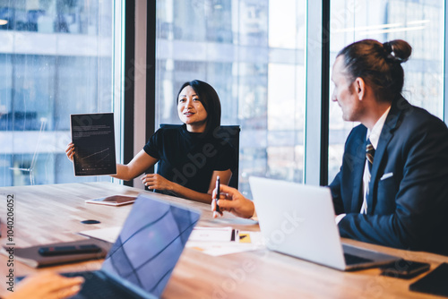 Elegant japanese woman in formal wear showing graphic with productive strategy for company working, group of business people collaborating on accounting in modern office sitting at desk with laptops