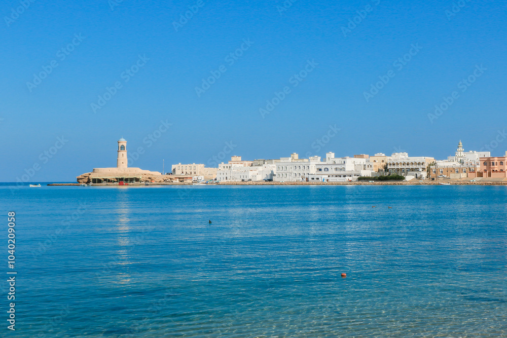 Coastal view of Sur Oman featuring historic buildings and a lighthouse along the shoreline under clear blue skies Coastal view of Sur Oman featuring historic buildings and a lighthouse along the shoreline under clear blue skies
