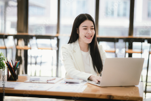 Beautiful Asian female accountant working with smartphone and laptop at her desk analyzing business reports and documents. Sending messages