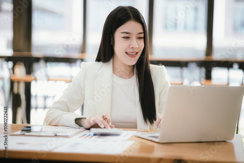 Beautiful Asian female accountant working with smartphone and laptop at her desk analyzing business reports and documents. Sending messages