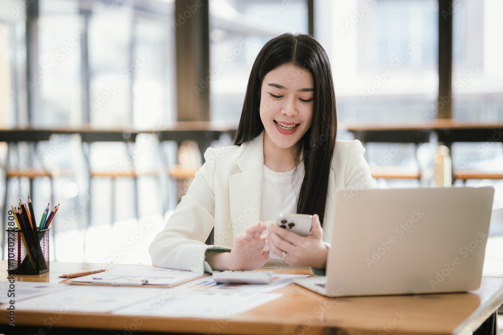 Beautiful Asian female accountant working with smartphone and laptop at her desk analyzing business reports and documents. Sending messages Beautiful Asian female accountant working with smartphone and laptop at her desk analyzing business reports and documents. Sending messages