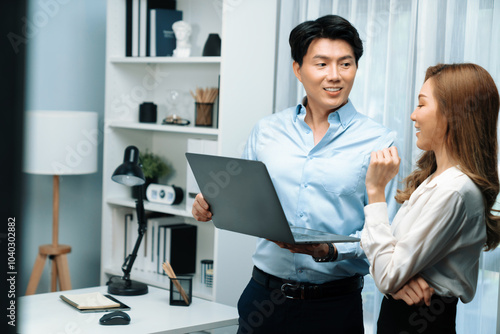 Smiling Asian coworkers standing while talking planning marketing report project on laptop casual day at modern office. Analyzing successful customer need in new social media online channel. Infobahn.