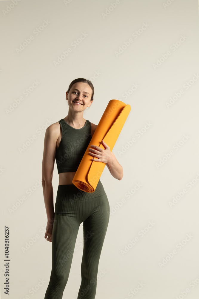 A young woman stands in a minimalist studio, holding an orange yoga mat. She is dressed in matching workout attire, radiating enthusiasm and readiness for exercise. A young woman stands in a minimalist studio, holding an orange yoga mat. She is dressed in matching workout attire, radiating enthusiasm and readiness for exercise.