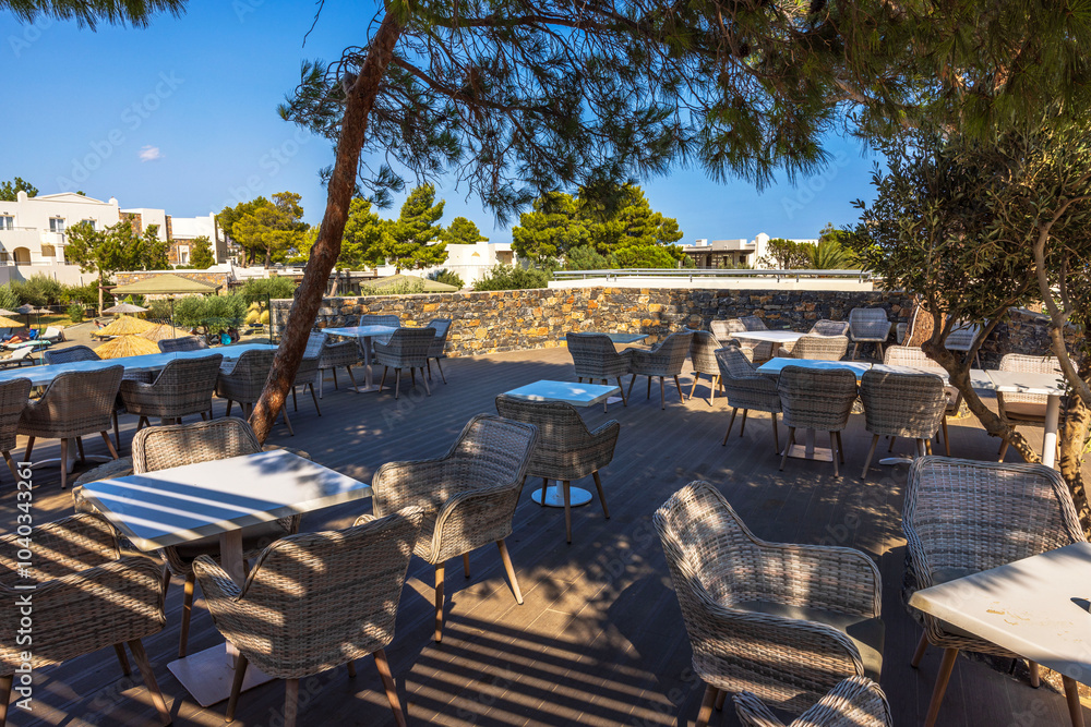Outdoor terrace with wicker chairs and tables under trees, with view of resort buildings and greenery. Crete. Greece. Outdoor terrace with wicker chairs and tables under trees, with view of resort buildings and greenery. Crete. Greece.