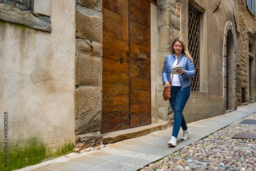 City break. Beautiful middle-aged woman in blue jacket and jeans walking on street, holding city map in old city of Bergamo Italy on autumn day. Front view. Season holidays.