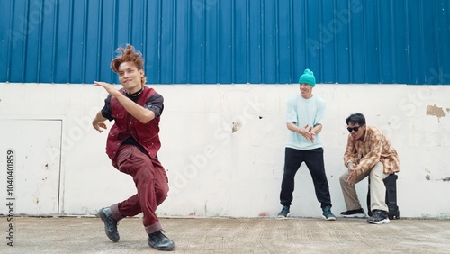 Young street dancer dancing in hip hop style with multicultural friends cheering behind at wall. Attractive handsome man stretch arms at street with blue background. Outdoor sport 2024. Hiphop. photo