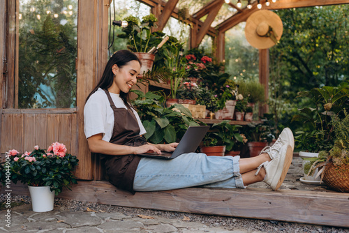 Side view of female in casualwear sitting on floor of glasshouse and holding computer on laps photo