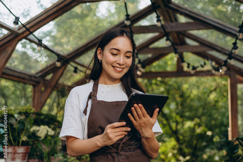 Portrait of smiling brunette lady standing at greenhouse and looking at digital tablet in hands