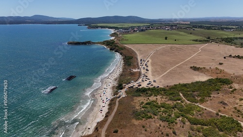 A stunning aerial view of a coastal landscape featuring a serene bay, vast green fields, and distant mountains under a clear blue sky. The image captures a peaceful seaside area with a few ships