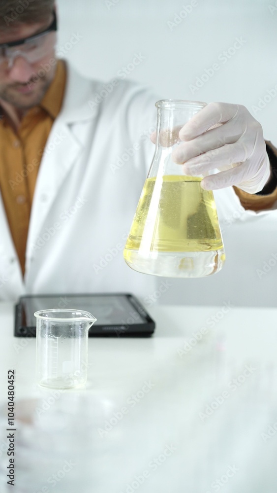 Man scientist wearing white protective gloves, and glasses is holding a yellow chemical solution inside an Erlenmeyer flask in a laboratory, vertical portrait Man scientist wearing white protective gloves, and glasses is holding a yellow chemical solution inside an Erlenmeyer flask in a laboratory, vertical portrait
