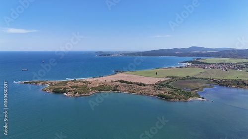A stunning aerial view of a coastal landscape featuring a serene bay, vast green fields, and distant mountains under a clear blue sky. The image captures a peaceful seaside area with a few ships