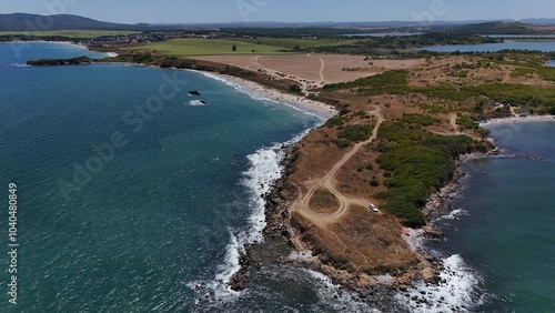 A stunning aerial view of a coastal landscape featuring a serene bay, vast green fields, and distant mountains under a clear blue sky. The image captures a peaceful seaside area with a few ships