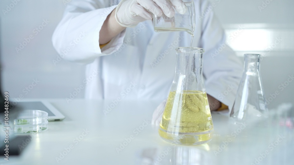 A scientist, wearing a lab coat and white protective gloves, is pouring a yellow oily liquid from one beaker to another in laboratory, close up. Medicine and science concept A scientist, wearing a lab coat and white protective gloves, is pouring a yellow oily liquid from one beaker to another in laboratory, close up. Medicine and science concept