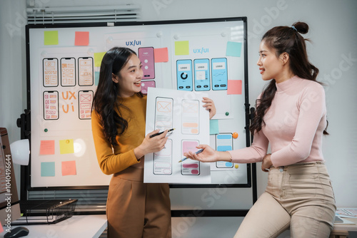 Two happy asian female uiux designers discussing over mobile application interface wireframe design together on whiteboard in modern office photo