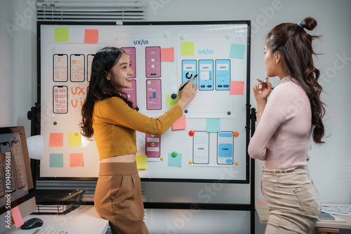 Two asian female ux ui designers are discussing over new mobile application interface design displayed on whiteboard, working together in office
