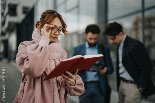 Focused female professional reading a notebook with male colleagues discussing in the background, portraying teamwork and collaboration outdoors.