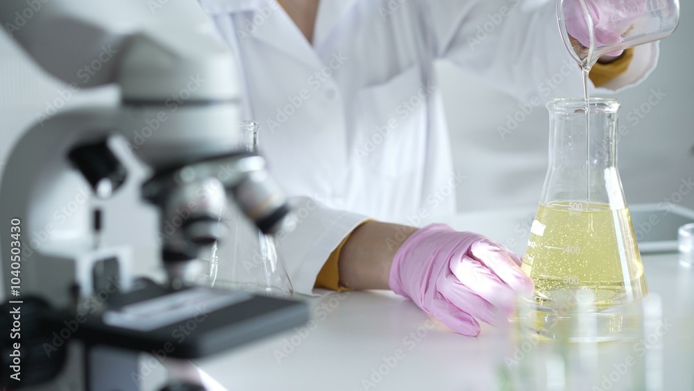 A scientist, wearing a lab coat and pink protective gloves, is pouring a yellow oily liquid from one beaker to another near microscope in laboratory, close up. Medicine and science A scientist, wearing a lab coat and pink protective gloves, is pouring a yellow oily liquid from one beaker to another near microscope in laboratory, close up. Medicine and science