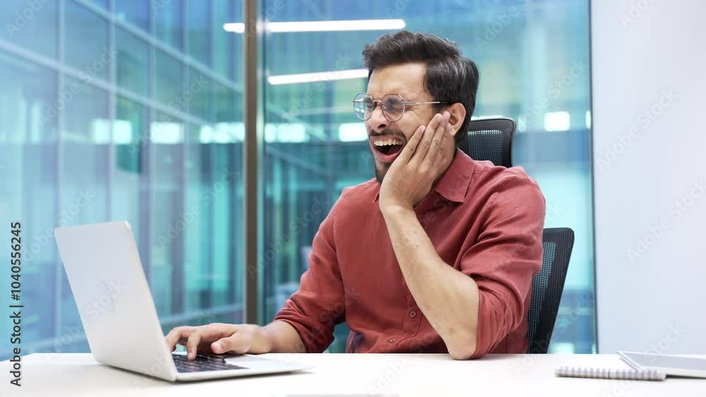 Upset businessman suffering from toothache working on laptop sitting at workplace in business office. Bearded worker holds his hand on his cheek, massaging the sore spot. He needs to see a dentist Upset businessman suffering from toothache working on laptop sitting at workplace in business office. Bearded worker holds his hand on his cheek, massaging the sore spot. He needs to see a dentist