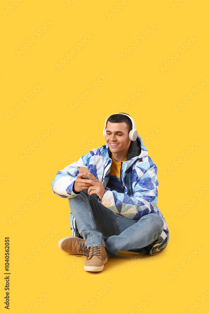 Young man in warm jacket with headphones using mobile phone on yellow background Young man in warm jacket with headphones using mobile phone on yellow background