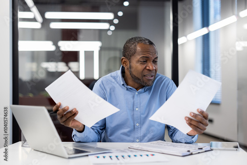 Mature businessman reviews documents at office desk with laptop and smartphone. African American professional shows frustration analyzing financial papers, embodying stress, focus, and determination.