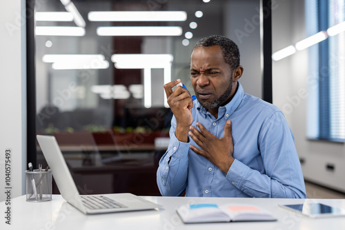 Mature african american businessman in office using inhaler. Man experiencing discomfort, holding chest while seated at work desk. Corporate setting with laptop and documents.