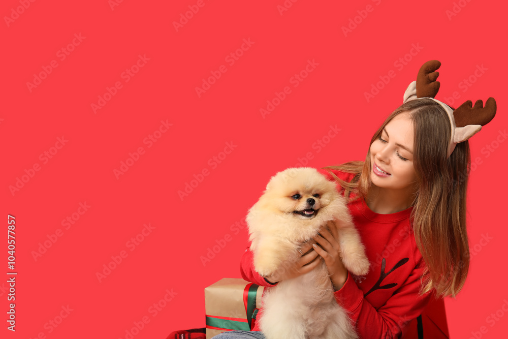 Young woman in reindeer horns and cute Pomeranian dog celebrating Christmas on red background Young woman in reindeer horns and cute Pomeranian dog celebrating Christmas on red background