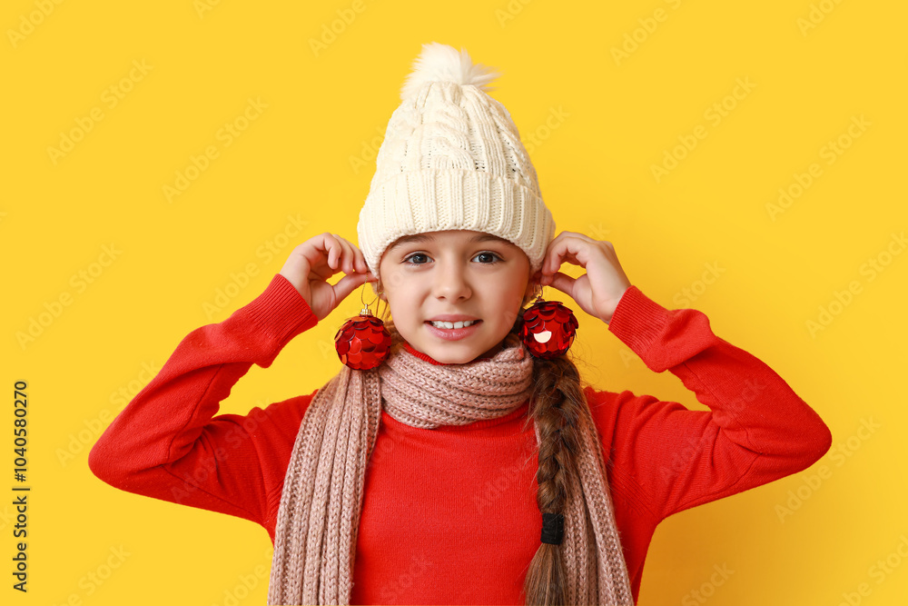 Funny little girl in warm hat and scarf with Christmas balls on yellow background Funny little girl in warm hat and scarf with Christmas balls on yellow background