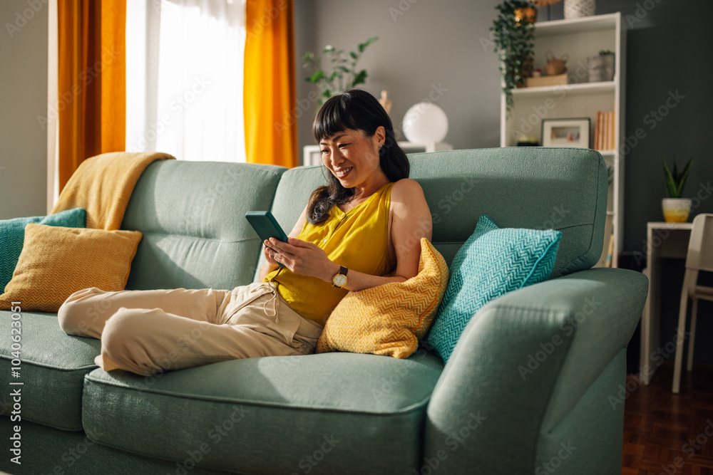 Obraz premium Asian woman in yellow top relaxes on teal sofa, using smartphone, cozy room.