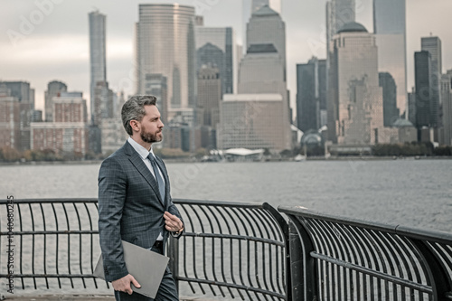 Business man in suit holding laptop on urban street in NYC. Successful business man outdoor. Middle aged business man standing outside office using laptop. Street portrait of mature businessman.