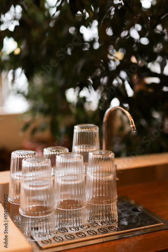 Stacked clear cups beside a modern water dispenser in a greenery-filled indoor setting during daylight hours