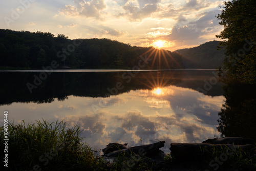 Sunrise over a calm small lake surrounded by a forest. Partly cloudy with sun rising over forested hills with a burst of sun rays and glowing clouds. Calm and peaceful photo