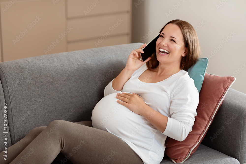 Fototapeta premium A happy pregnant woman is sitting on a couch, laughing joyfully as she speaks on her phone. She holds her belly comfortably, enjoying a relaxed moment at home in the afternoon.